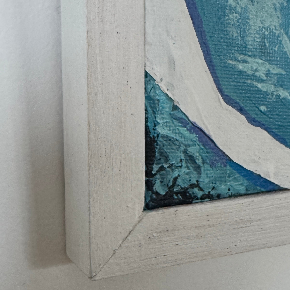 Close-up of a framed blue painting corner showing a white textured frame and blue, teal, and white painted canvas detail.