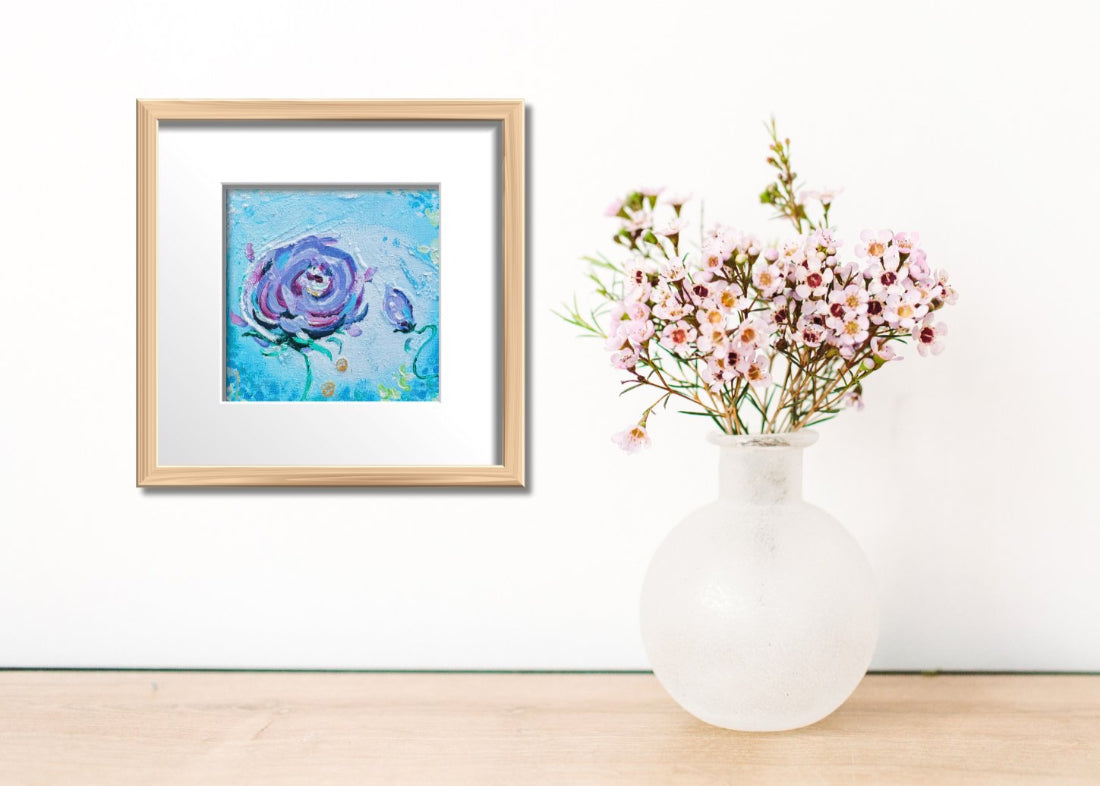 Framed artwork of a blue flower on a wall next to a white vase with flowers on a wooden surface.