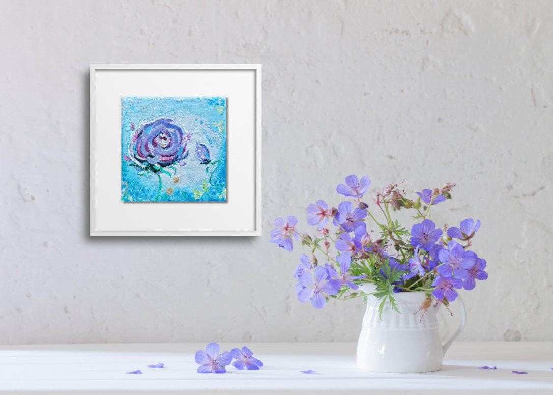 Framed artwork of a blue rose on a wall with a vase of purple flowers below.