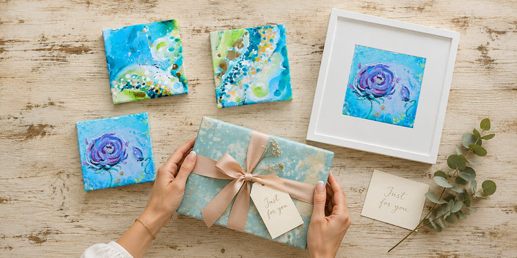 Abstract art gifts on a wooden table — small blue and green canvases, a framed purple rose print, and a wrapped gift with a "Just for you" tag