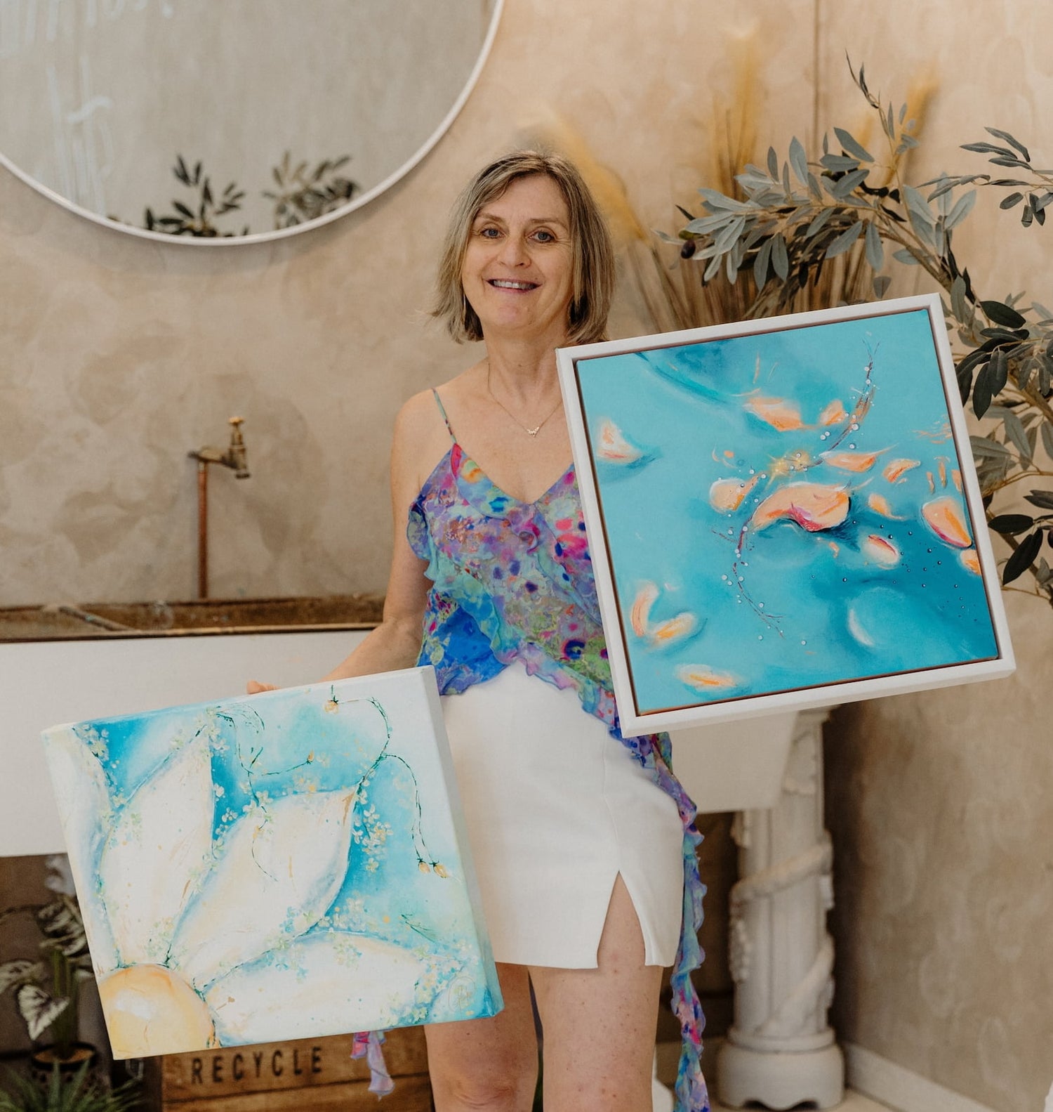 A smiling woman stands indoors holding two framed abstract artworks. One painting is predominantly turquoise blue with soft peach and cream brushstrokes, and the other shows large white and light blue petal-like forms with gold accents.