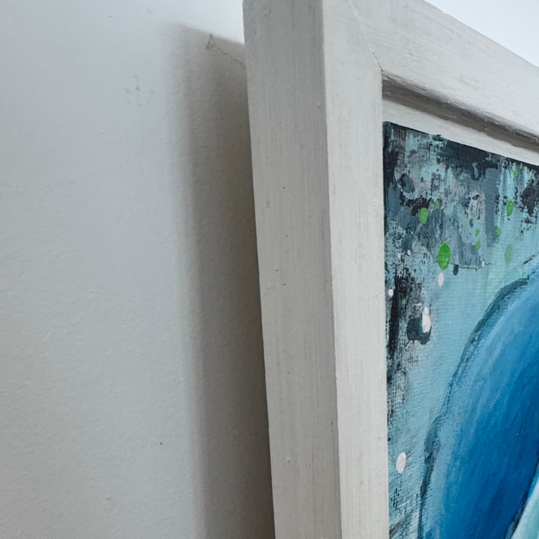 Close-up of framed abstract painting Beautiful Mess showing a white wooden frame corner and textured blue, gray, black, and green paint on a light wall background.