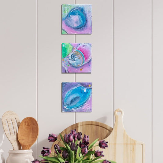 Three small abstract paintings on a white wall above a vase of purple tulips and wooden cutting board.