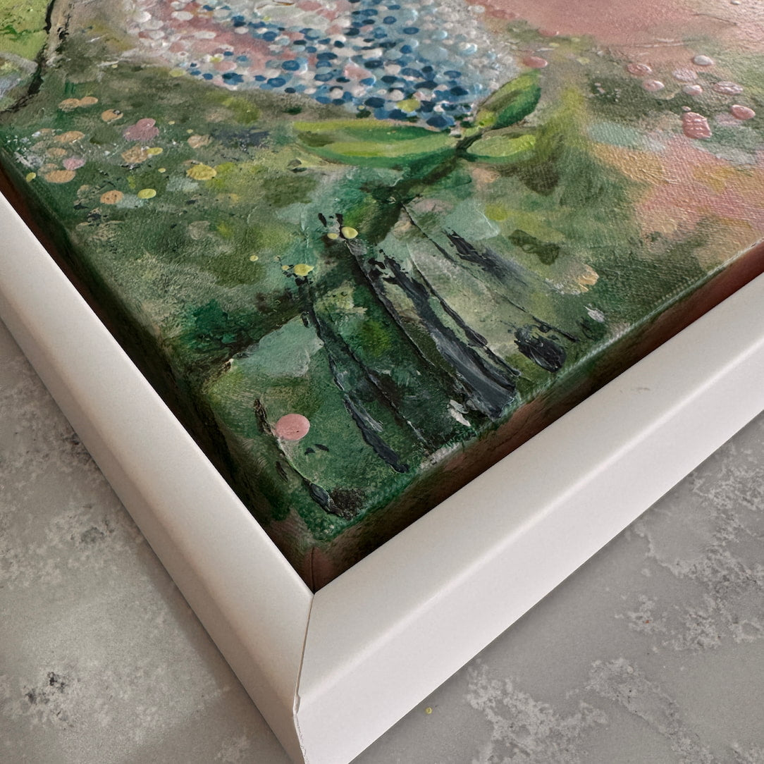 A close-up corner view of a framed artwork showing textured green foliage, blue and pink floral details, and a white frame on a gray floor.