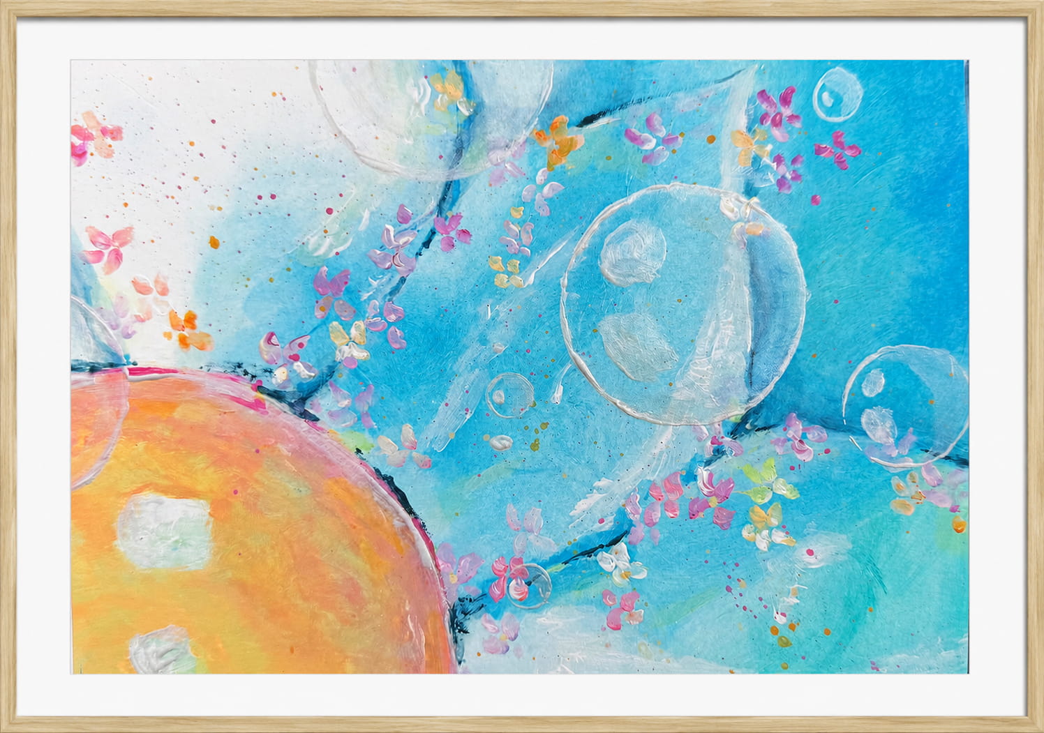 A framed abstract art print featuring a vibrant blue background with watercolor-style splashes, delicate pink and yellow flowers, translucent bubble shapes, and a large warm orange-yellow form in the lower left, all enclosed in a light natural wood frame with a white mat.