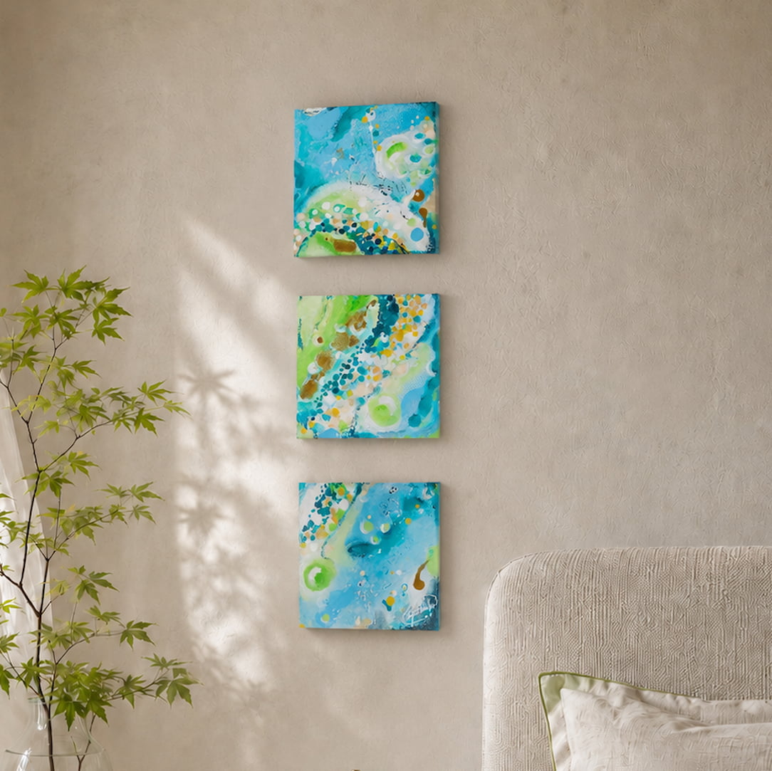 Three square wall art set with abstract blue and green designs hang vertically on a sunlit wall.