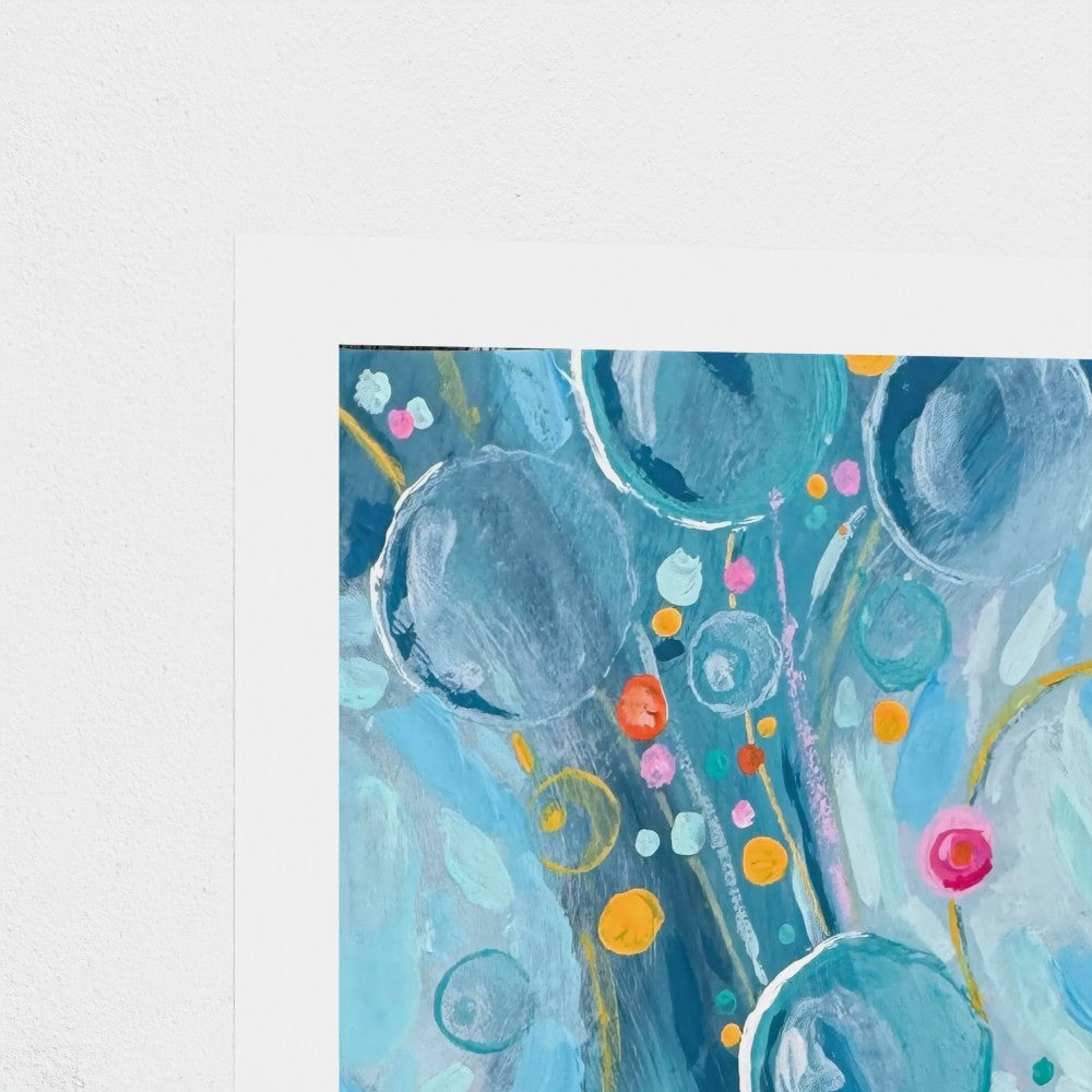 Close-up of an abstract art print with blue and teal bubble-like circles with colorful painted round shapes on a white paper border, shown against a light gray wall background.