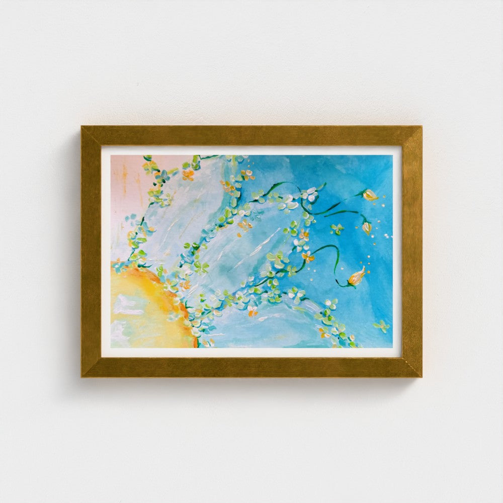 A framed floral wall art print with a bright blue background with white brushstrokes, delicate green and yellow floral branches, and a soft yellow sun shape, housed in a textured gold frame and mounted on a white wall.
