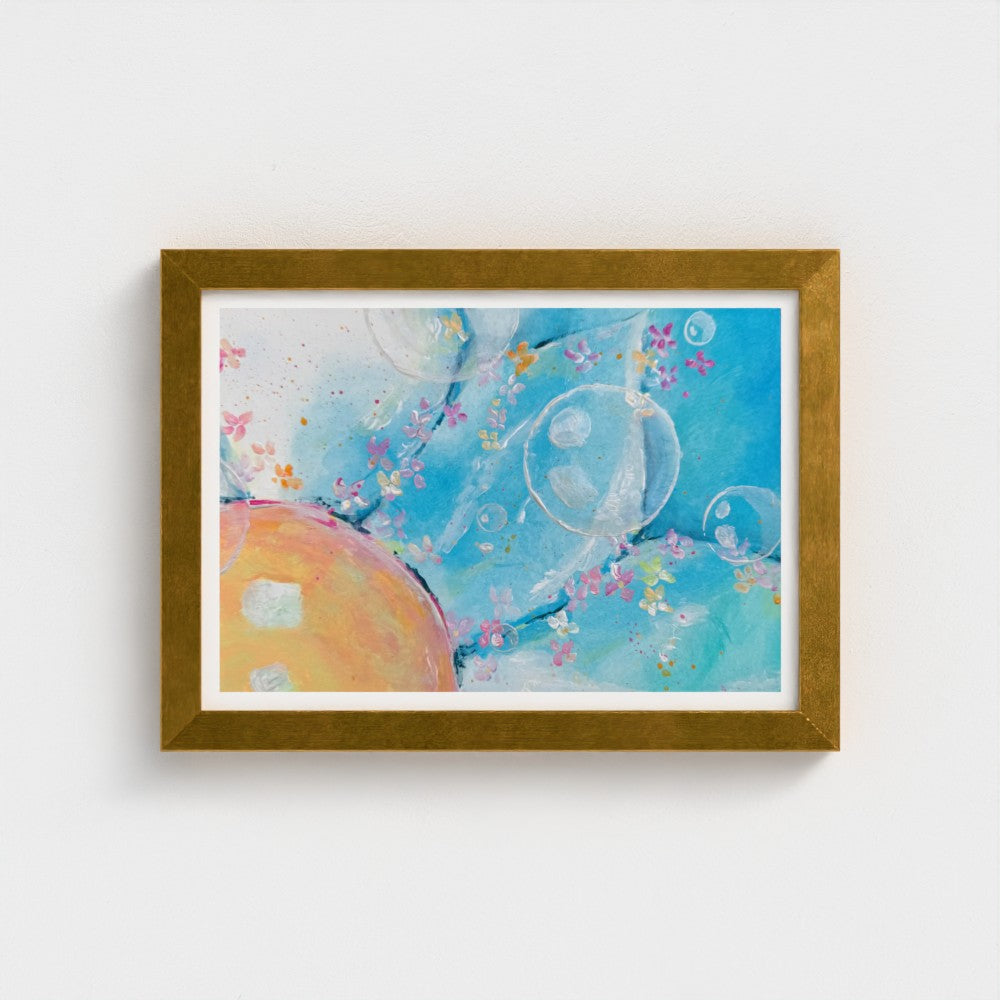 A framed abstract art print featuring a light blue background with watercolor-like bubbles, small pink and yellow flowers, and a peach-colored oval shape, set in a textured gold frame against a white wall.