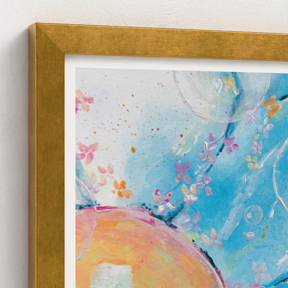 A close-up of a framed abstract watercolor art print with a textured gold frame, showing blue, pink, orange, and white painted details against a white wall.