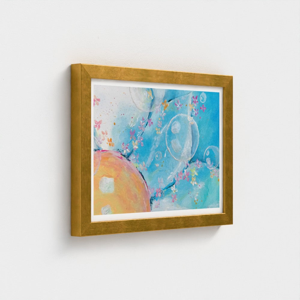 Framed abstract wall art hanging on a light gray wall. The artwork features a blue background with soft bubble-like forms, pastel floral details, and a peach-colored circular shape, surrounded by a white mat and a metallic gold frame.