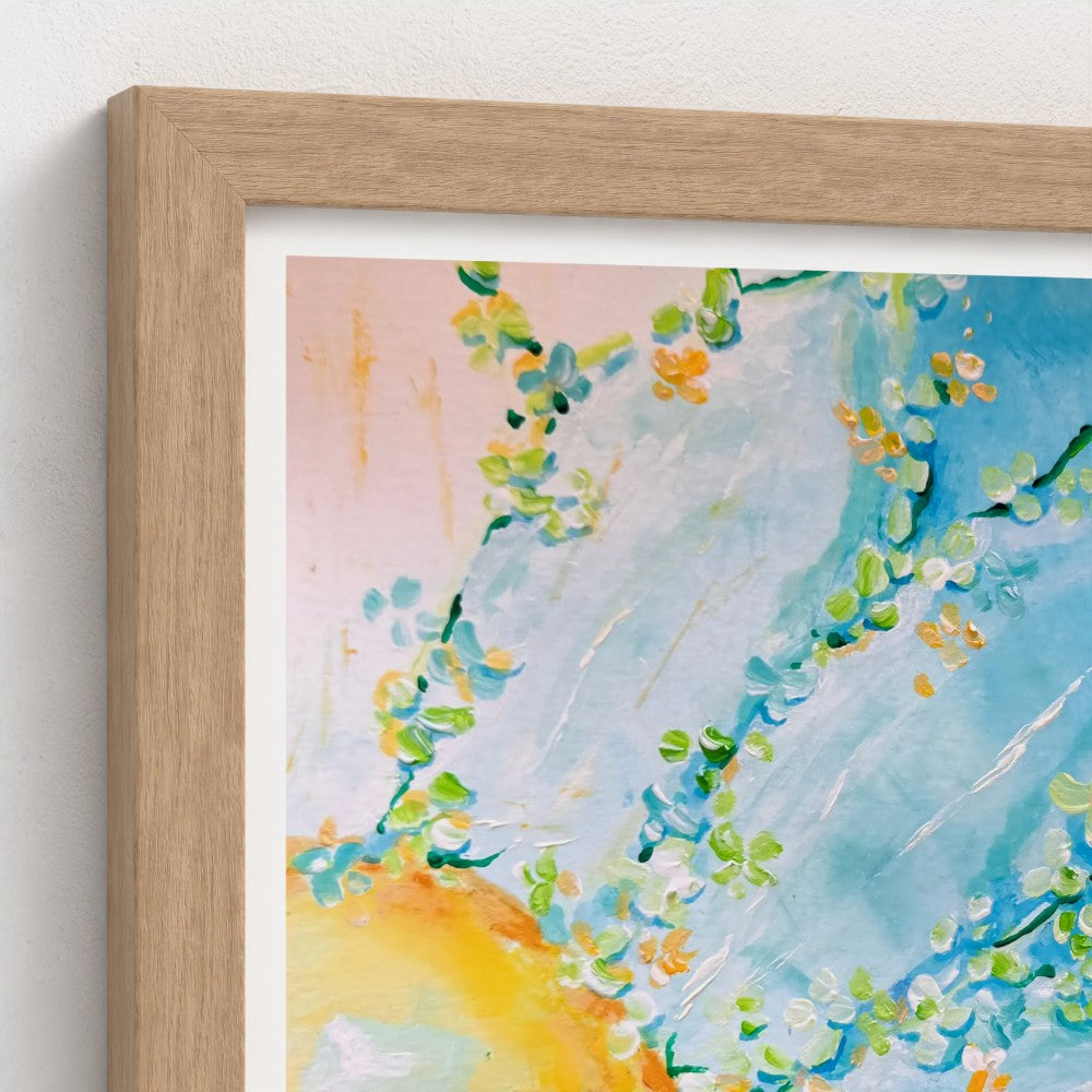 A close-up of a framed art print featuring a watercolor painting of blossoming branches with green and yellow flowers against a blue and pink sky, set in a light oak frame.
