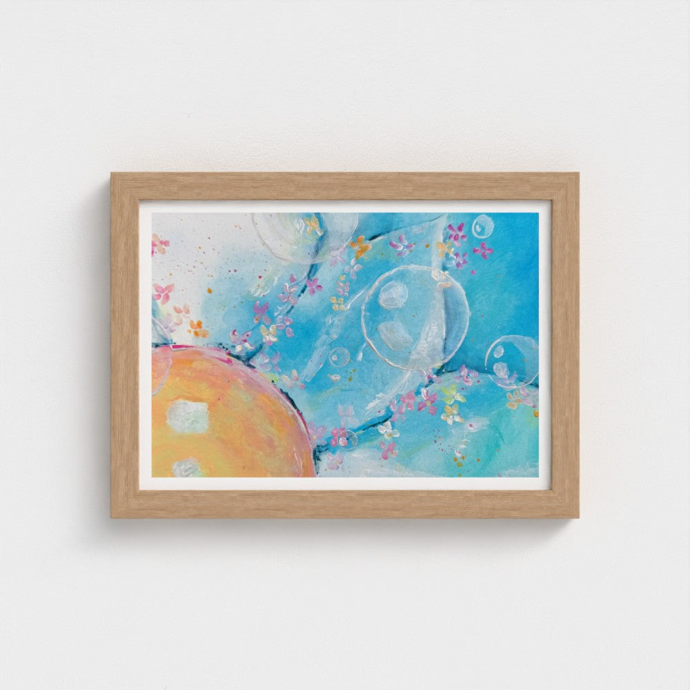 A framed abstract art print featuring a light blue background with watercolor-like splashes, translucent bubble shapes, and a warm peach-orange circular form, set in a natural wood frame against a white wall.