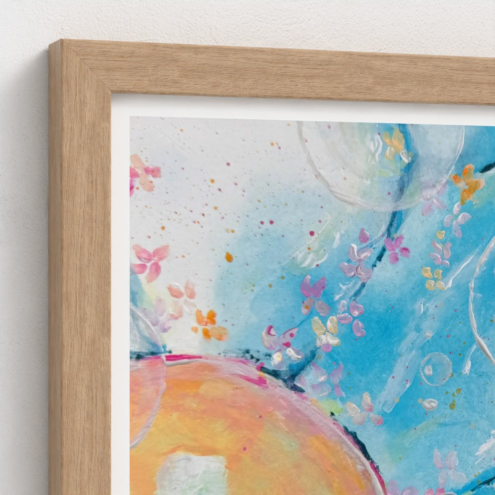 A close-up of a framed abstract art print featuring a light wood frame, white mat, and a colorful painting with blue, pink, orange, and yellow watercolor textures on a white wall.