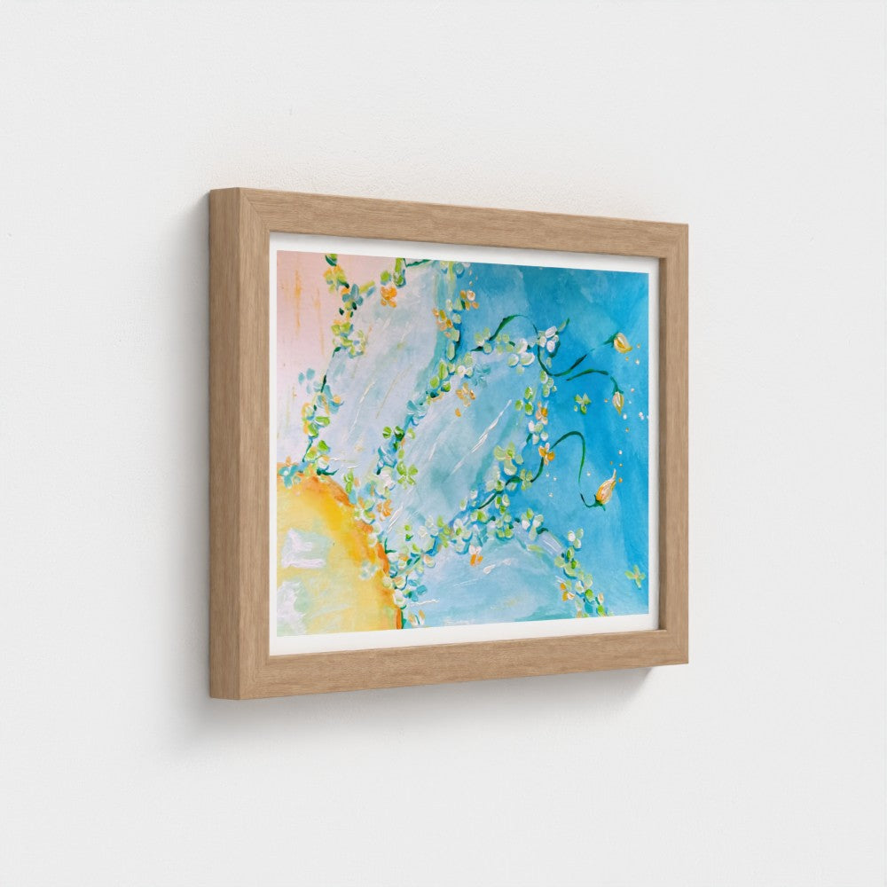 Framed abstract floral art print with blue, white, yellow, and soft peach tones, shown in a light oak wood frame against a white wall.