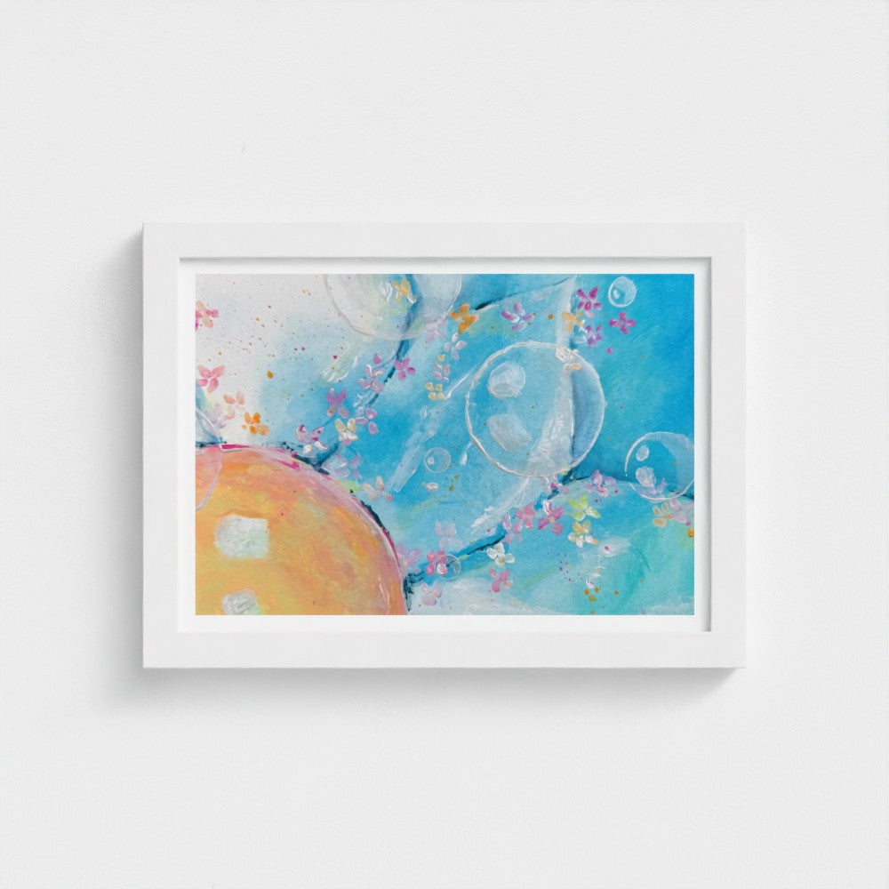 A framed abstract art print featuring a light blue background with watercolor textures, translucent bubble-like shapes, small pink and yellow floral accents, and a large peach-orange oval shape in the lower left. The print is housed in a clean white frame and displayed against a neutral light gray wall.