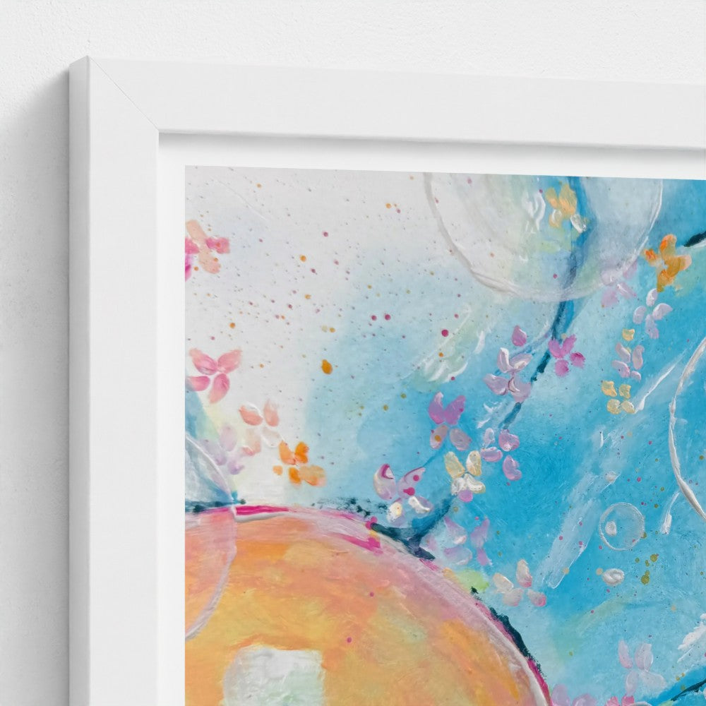 A close-up of a framed abstract art print with a white frame, showing a watercolor-style design in shades of blue, orange, pink, and yellow.
