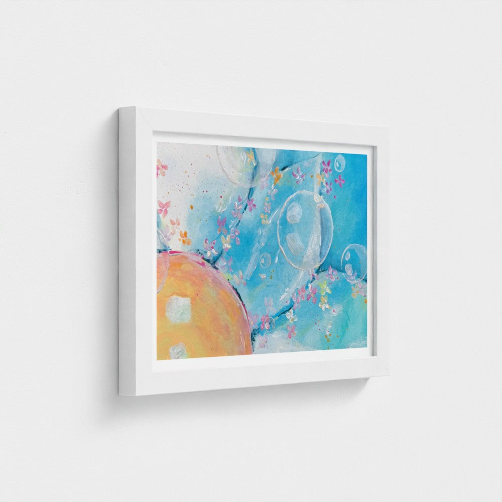 A framed abstract art print featuring a vibrant blue background with floating bubble-like shapes and a large warm orange-yellow form, presented in a white frame against a light gray wall.