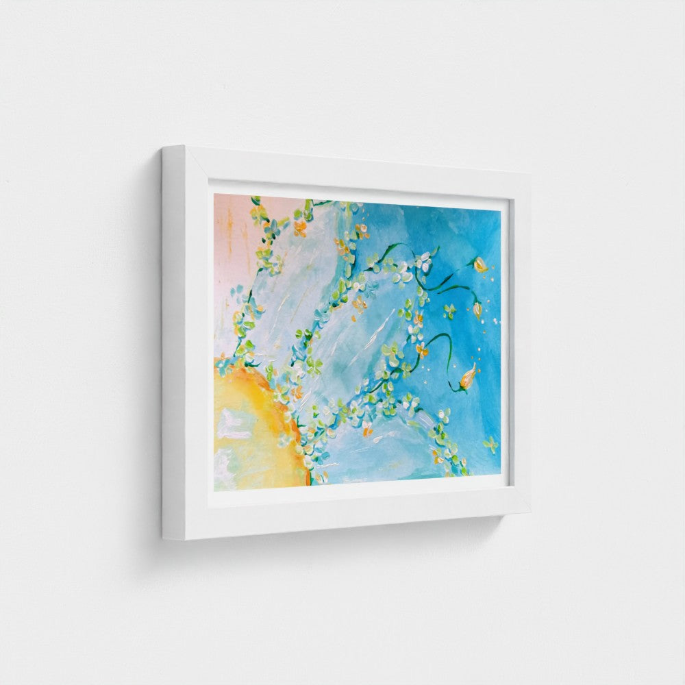 A framed abstract fine art paper print featuring a vibrant blue background with white blossoms, green leaves, and a warm yellow-orange glow in the lower left, presented in a white frame against a light gray wall.